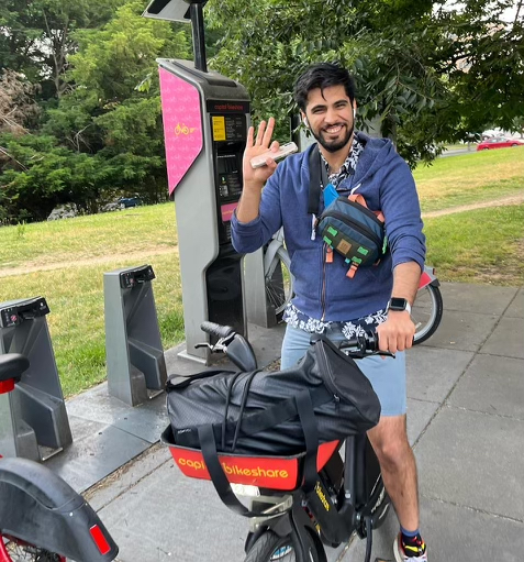 Himanshu Ahuja sitting on a rental bicycle, wearing a blue hoodie and shorts, and smiling and waving at the camera.